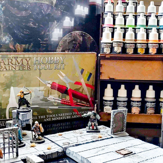 Image of painted miniatures arranged on a grey stone ground set piece. Behind them is the Army Painter Hobby Tool Kit box which is next to four rows of miniature paints on a stacked wooden rack. Image exclusively on Kaioss Games.