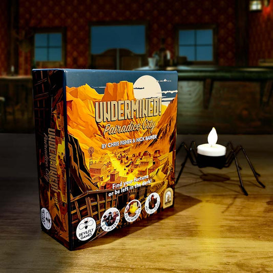 Image of Undermined! Pairadice City on a table with a small candle behind it. Behind the table is a very dark, old western saloon-esque room with dark blue in the windows. Exclusively on the Kaioss Games blog..