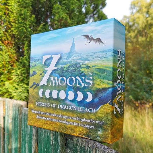 Image of the 7 Moons (Deluxe) board game box on top of a green fence with green trees and grass in the background. Image exclusively on the Kaioss Games blog