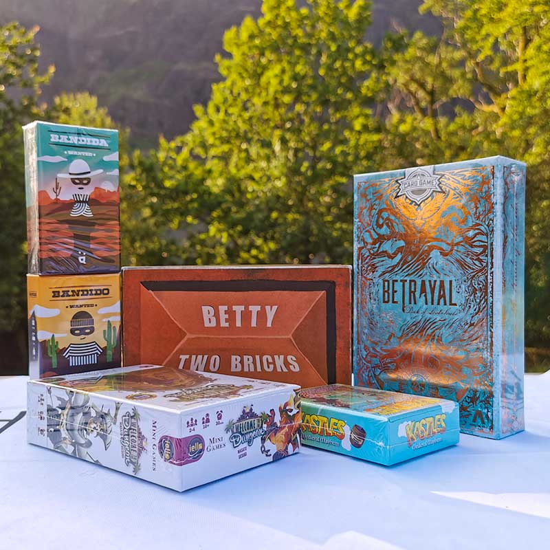 Image of card games on a wooden table. Behind the table are green bathed in sunlight. The card games from left to right are Bandido and Bandida, Betty Two Bricks, Welcome to the Dungeon, Kastles and Betrayal Deck of Lost Souls