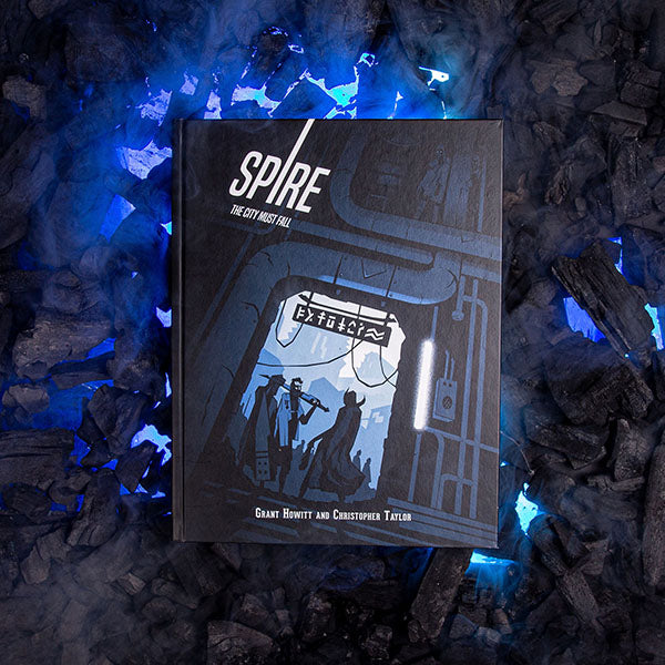 Image of Spire: Core Rule Book, 5th Anniversary Edition RPG book on top of a pile of burn wood with purple light shining through the wood pieces. This game by Rowan, Rook & Decard is available at indie online boardgames and card games retailer Kaioss Games