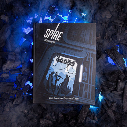 Image of Spire: Core Rule Book, 5th Anniversary Edition RPG book on top of a pile of burn wood with purple light shining through the wood pieces. This game by Rowan, Rook & Decard is available at indie online boardgames and card games retailer Kaioss Games